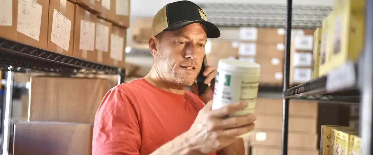 A man checking a product while speaking on the phone beside stocked shelves.